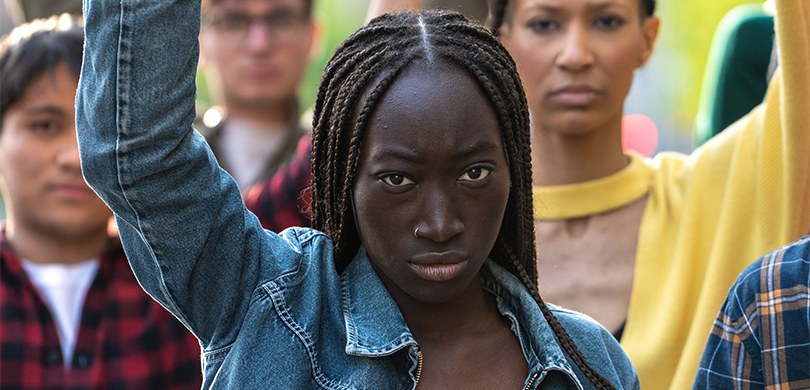 Foto em que aparece em primeiro plano uma mulher negra, de cabelos trançados, em pé, com o braço...